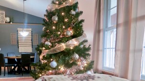 Un salon chaleureux avec un sapin de Noël décoré, un coussin à motifs et un coin repas comprenant une table et des chaises.