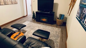 Sala de estar com lareira, televisão e área de estar com sofás e pufes de couro.