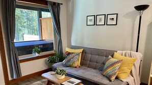 Sala de estar com um sofá cinzento, uma mesa de centro de madeira e uma janela com cortinas. Há três quadros na parede e um abajur.