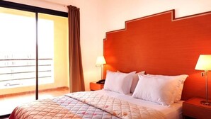 Habitación de hotel con una cama grande, cabecera de madera, dos mesitas de noche con lámparas y vistas a un balcón con barandilla.