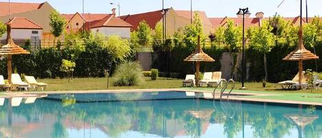 A swimming pool with lounge chairs and umbrellas, surrounded by greenery and buildings with red roofs.