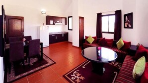 Ein Wohnzimmer mit rundem Couchtisch, einer Couch mit roten Kissen, einem Essbereich mit Tisch und Stühlen sowie einer Küche im Hintergrund.