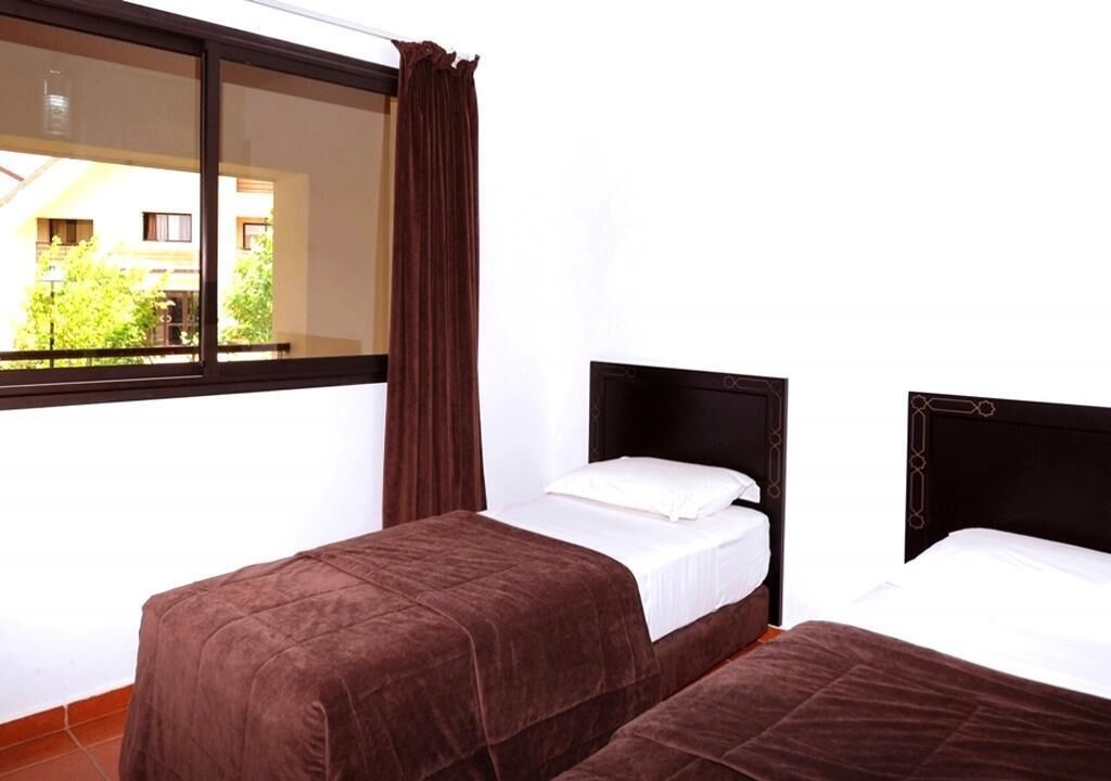 Une chambre avec deux lits, une fenêtre avec des rideaux et une vue sur les bâtiments et les arbres à l’extérieur.
