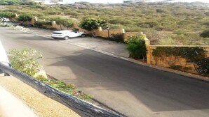 A residential street with a white car parked on the side, surrounded by greenery and a stone wall. - Residence Les Jardins de La Lagune 2275 (Oualidia)