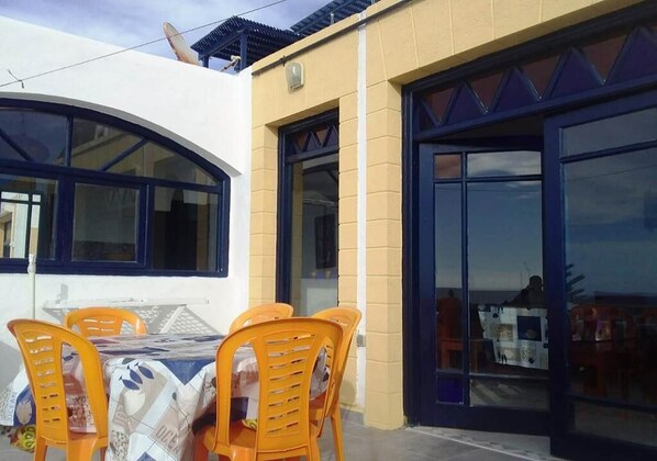 An outdoor dining area with a table and chairs, a building with large windows and a door, and a clear sky.