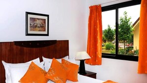 Ein Hotelzimmer mit einem Bett, orangefarbenen Kissen, einem gerahmten Bild, einem Fenster mit orangefarbenen Vorhängen und Blick auf Grünfläche vor dem Fenster.