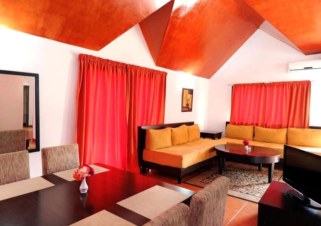 Un salon moderne avec un canapé, une table basse et un téléviseur. On y trouve également une table à manger avec des chaises et un vase de fleurs. Des rideaux rouges sont visibles aux fenêtres.