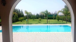 Une vue à travers une porte en arc donnant sur une piscine, avec un jardin aménagé et des arbres en arrière-plan.