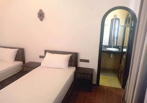 A hotel room with two beds, a small bedside table, a wall-mounted light, and a bathroom visible through an archway.