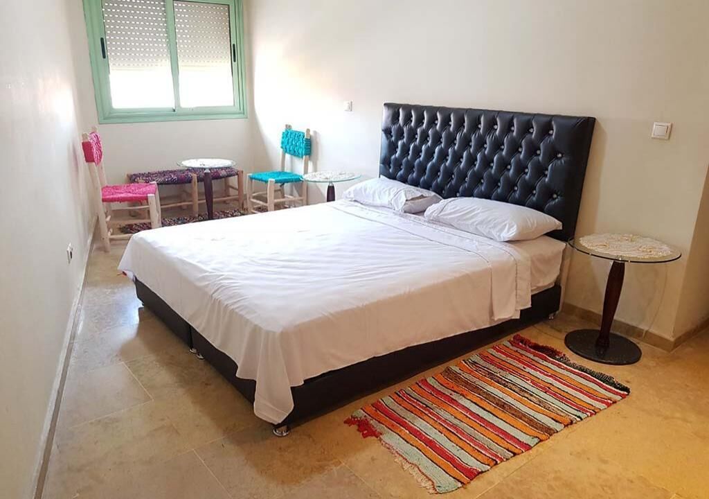 Une chambre à coucher avec un grand lit, une petite table ronde, une chaise et une fenêtre dotée d’un cadre vert.