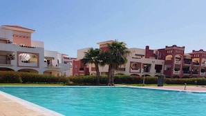 A swimming pool with a clear blue water, surrounded by multi-story buildings with balconies and a palm tree. - Very nice apartment El Waha Saidia 4597 (Saidia)