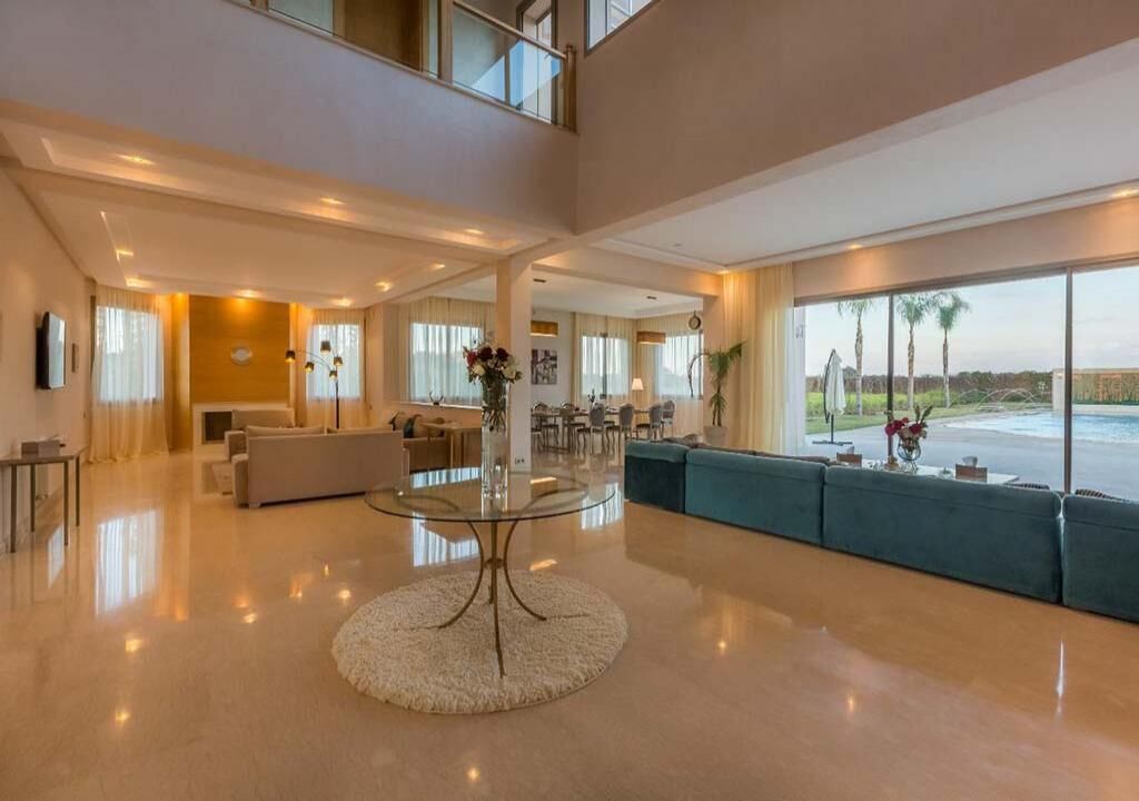 Un salón amplio y moderno con techo alto, una mesa de centro redonda de vidrio y un gran ventanal con vistas a palmeras.