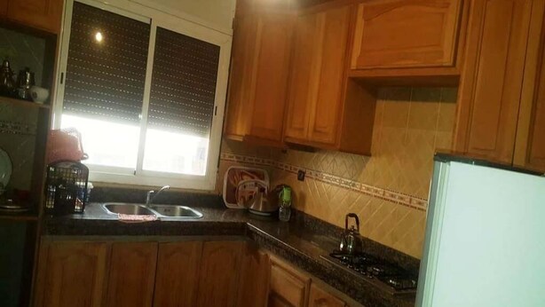 Una cocina con armarios de madera, un lavamanos de acero inoxidable, una tetera y una ventana con persianas.