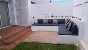 Ein moderner Sitzbereich im Freien mit einer eingebauten Couch und Kissen, einer kleinen Stufe zum Eingang und einem Rasenstück.