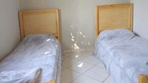 Ein Zimmer mit zwei Holzbetten vor einer schlichten Wand, wobei Sonnenlicht Schatten auf den Boden wirft.