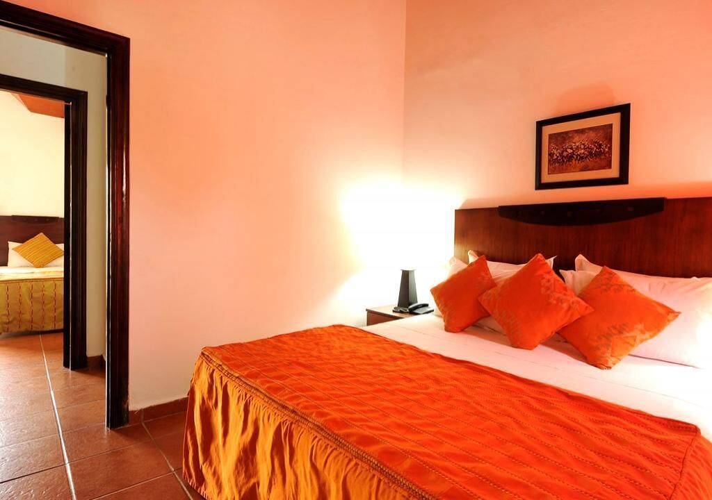 A hotel room with a bed, orange bedding, a nightstand with a lamp, and a framed picture on the wall.