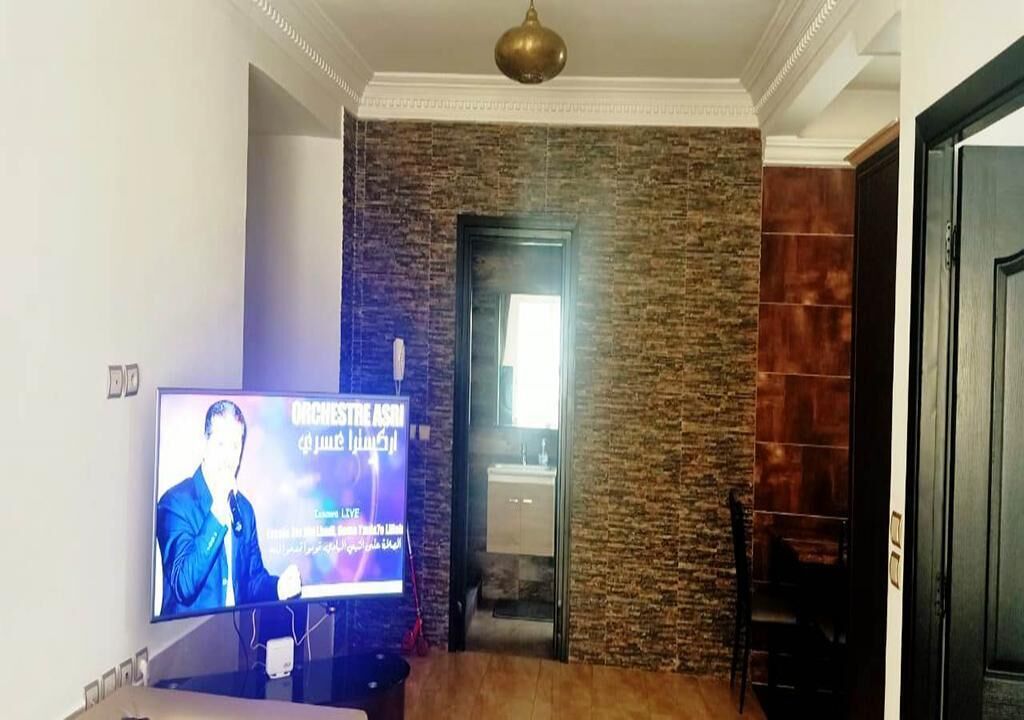 Habitación con televisión mostrando un evento en vivo, pared de piedra, una puerta y una silla.
