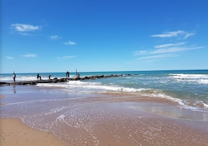 A beach with clear skies, calm waters, and a rocky outcrop where people are standing.