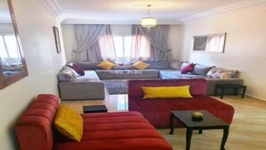 A living room with a red sofa, a grey sofa, and a red armchair. There is a small table and a window with curtains. - Pleasant apartment in Jawharat Mogador 4729 (Essaouira)