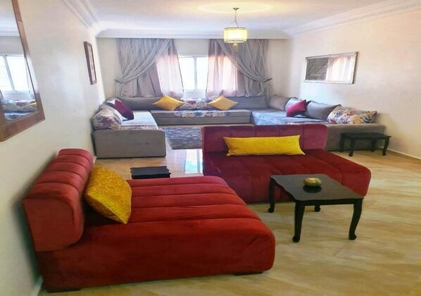 A living room with a red sofa, a grey sofa, and a red armchair. There is a small table and a window with curtains.