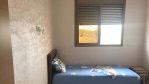A bedroom with a single bed, a window with blinds, a wall socket, and a small bedside table.