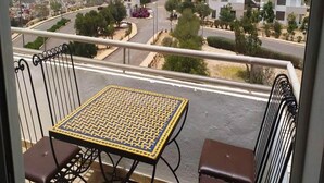 Un balcon avec une table et deux chaises, donnant sur une rue bordée d’immeubles et d’espaces verts.
