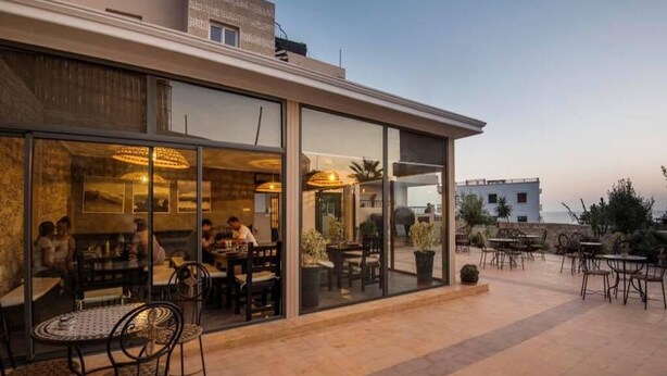 An outdoor dining area with tables and chairs, a glass wall separating the dining area from the interior, and a view of the cityscape in the background. - Moroccan Surf Adventures 5185 (Imi Ouaddar)