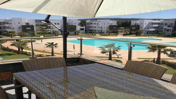 Zona junto a la piscina con mesa y sillas, una sombrilla grande, y vista de una piscina y edificios al fondo.