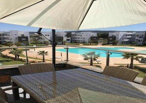 Een zwembadterras met een tafel en stoelen, een grote parasol en zicht op een zwembad en gebouwen op de achtergrond.