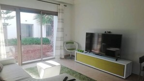 Una sala moderna con un sofá, un televisor sobre un mueble blanco y amarillo, una silla y un ventanal corredizo que da a un jardín.