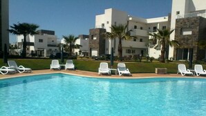 En swimmingpool omgiven av vita solstolar, med moderna byggnader och palmer i bakgrunden.