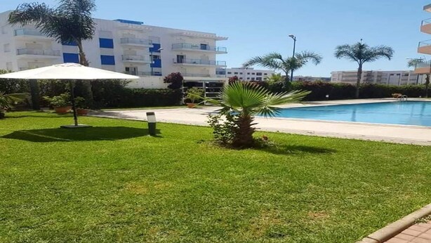 A pool area with a well-maintained lawn, a palm tree, and a building with balconies in the background.