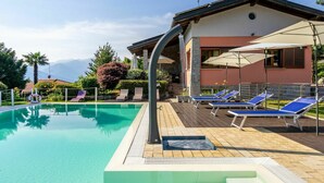 En swimmingpool med liggestole og parasoller, omgivet af en anlagt have og en bygning med en stenfacade.