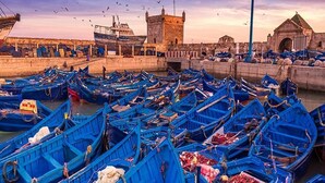 Ein Hafen voller blauer Boote, ein steinernes Turmgebäude und ein Haus mit spitzer Dachform.