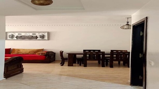 Ein geräumiger Wohnbereich mit einem roten Sofa, einem Esstisch mit Stühlen, einer Deckenleuchte und einem gerahmten Bild an der Wand.