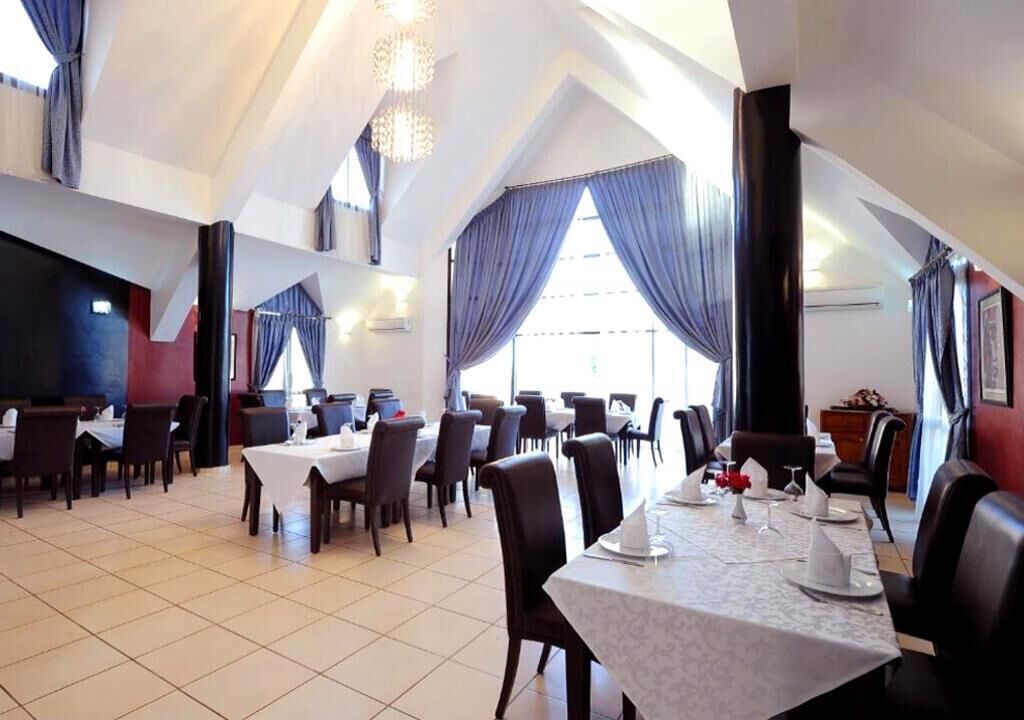 Un comedor amplio con varias mesas y sillas, ventanas grandes con cortinas azules y un techo alto con candelabros.