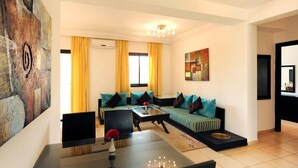 Ein Wohnzimmer mit einer Sofagarnitur, einem Couchtisch und einem Fernseher. An der Wand hängt ein Gemälde, und ein Fenster mit Vorhängen ist ebenfalls zu sehen.