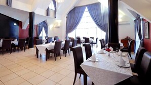 Un'ampia sala da pranzo con diversi tavoli e sedie, grandi finestre con tende blu e un soffitto alto con lampadari.