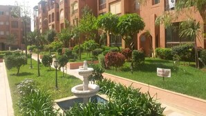 Un patio con una fuente, rodeado de follaje y un edificio de varios pisos con balcones.