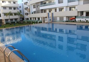 A swimming pool with a metal ladder, surrounded by modern buildings with balconies and windows. - Pleasant apartment in Paradise Beach pool view 4848 (El Mansouria)