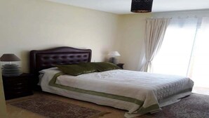 A bedroom with a bed, two bedside tables, a nightstand, a window with curtains, and a hanging light fixture. - Apartment with pool view in Kalaat Al Mansourya 4850 (El Mansouria)