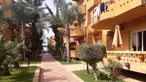 A pathway leading to a building with balconies, surrounded by greenery and palm trees. - Apartment with pool view in Kalaat Al Mansourya 4850 (El Mansouria)