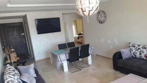 A living room with a sofa, a dining table with chairs, a TV mounted on the wall, and a clock. - Apartment with pool view in Kalaat Al Mansourya 4850 (El Mansouria)