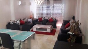 A modern living room with a dining table, two sofas with red cushions, a coffee table, and a decorative plant. - Apartment with pool view in Kalaat Al Mansourya 4850 (El Mansouria)
