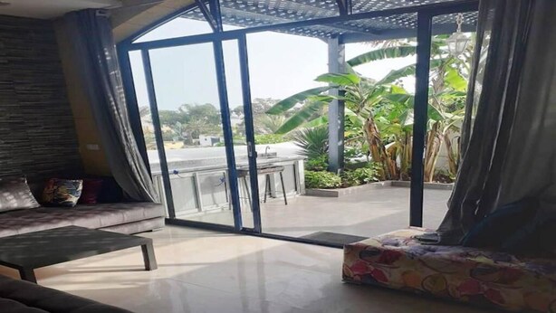 Una sala de estar moderna con un sofá, una mesa de centro y un reposapiés estampado. Un amplio ventanal ofrece vistas a un patio con plantas y un comedor.
