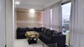 A room with a sectional sofa, a small table with a tablecloth, a window with curtains, and a view of the outdoors.