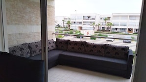 A balcony with a dark-colored sofa and patterned cushions, overlooking a street with buildings and palm trees.