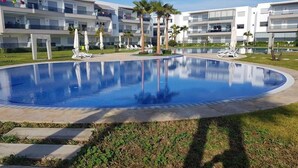 A swimming pool surrounded by a grassy area, with white buildings in the background. - Pleasant Apartment in Blanca Beach 4873 (Sidi Rahal)
