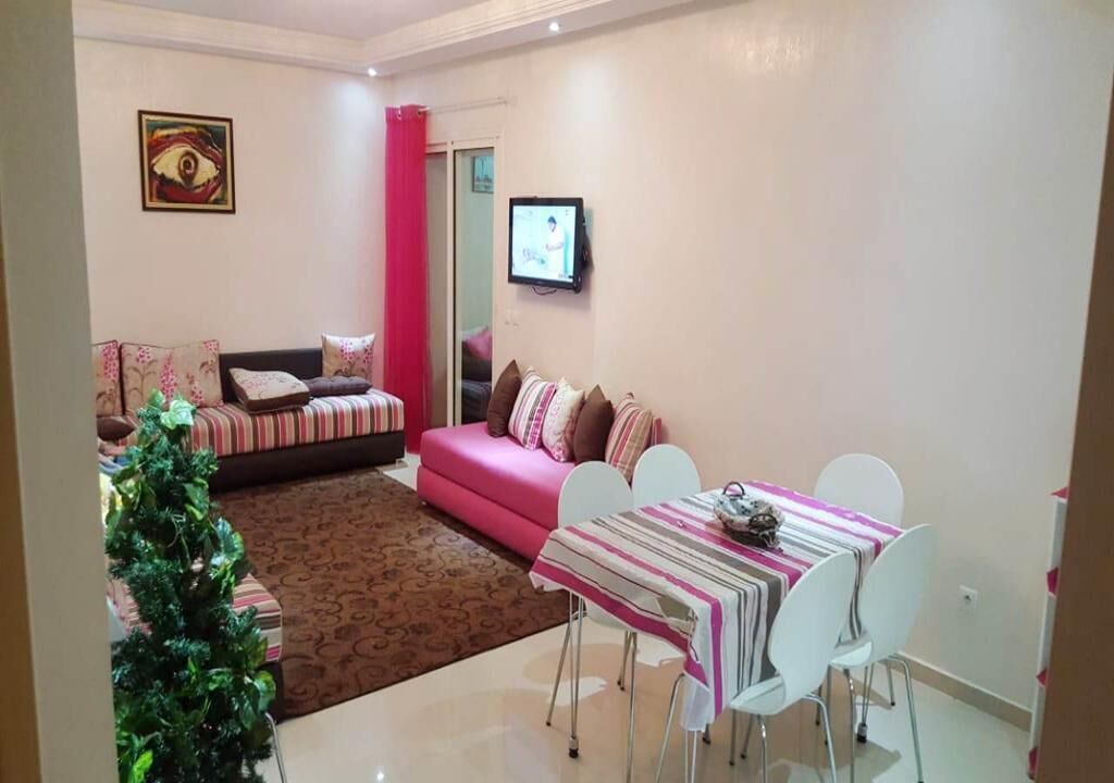 Un salon avec un canapé rose, une table à manger recouverte d’une nappe à rayures et un petit sapin de Noël.
