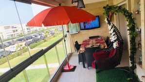 Un balcón con una sombrilla roja, una zona de comedor con mantel estampado, y vistas a coches aparcados y vegetación.